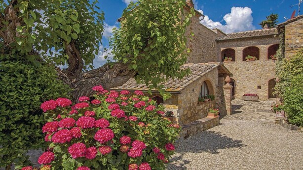 Un bâtiment en pierre doté d’une cour intérieure, entouré d’une végétation luxuriante et de fleurs roses éclatantes.