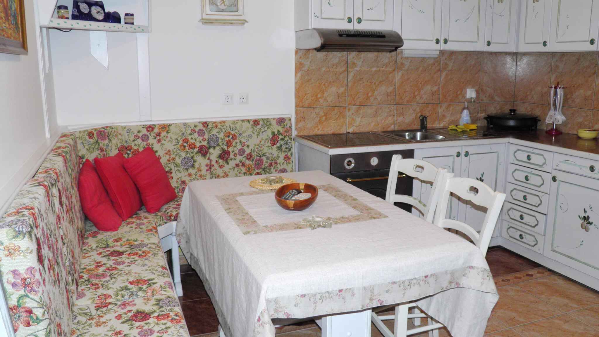 Un accogliente soggiorno con un divano floreale, un tavolo da pranzo e un angolo cottura con pensili bianchi e piastrelle a vista.