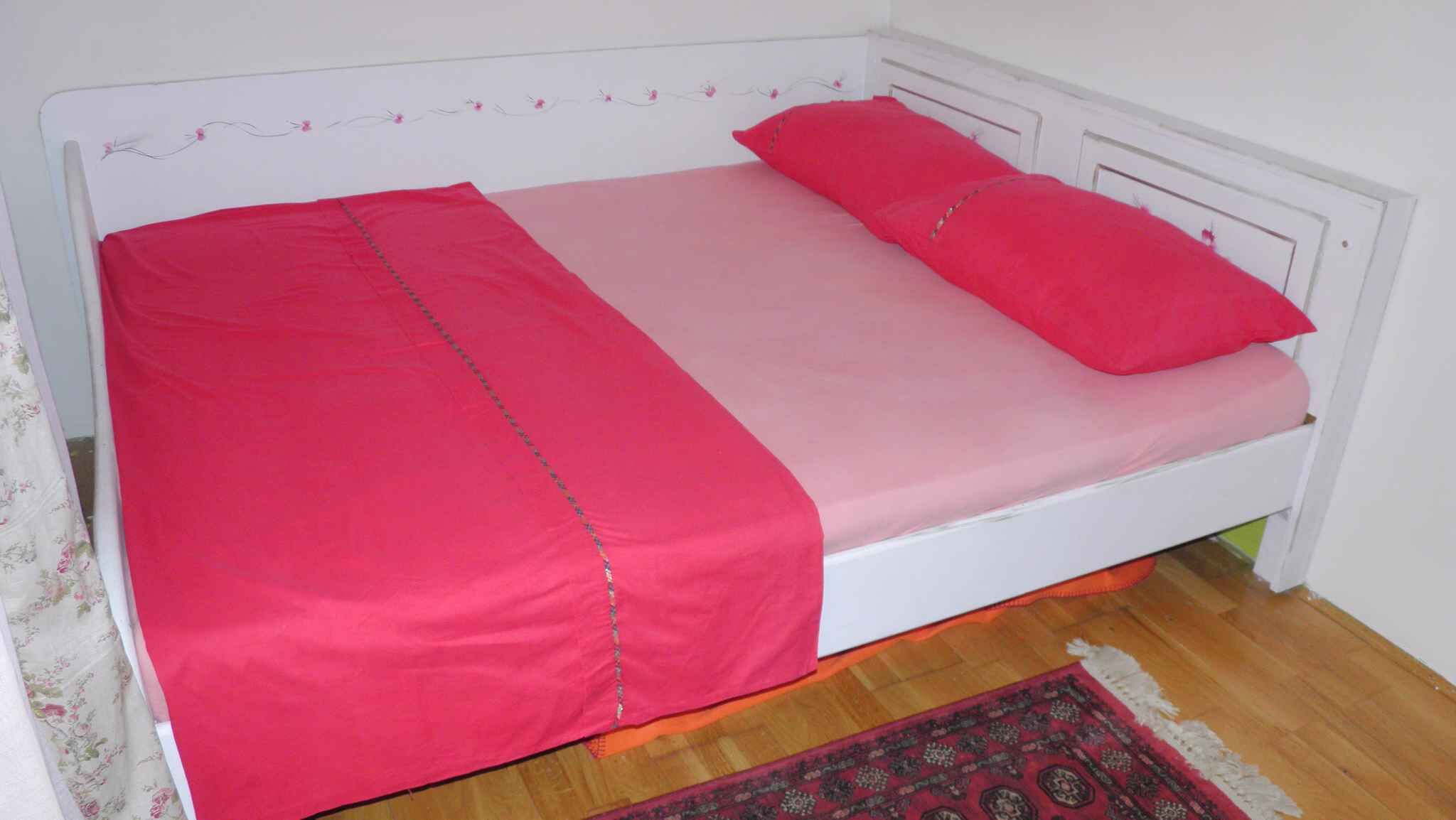 Habitación individual con una cama que tiene cabecera blanca, ropa de cama rosa y una alfombra estampada sobre piso de madera.