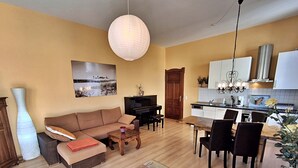 Ein Wohnzimmer mit Sofa, Couchtisch und Fernseher, ein Essbereich mit Stühlen und Tisch sowie eine Küche mit Schränken und Spüle.