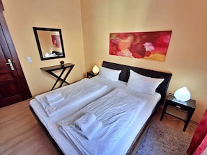 A neatly made bed with white linens and towels, a bedside lamp, a small table with a vase of flowers, a wall-mounted mirror, and a large abstract painting.