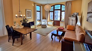Ein geräumiges Wohnzimmer mit einem Holzesstisch, schwarzen Stühlen, einem braunen Sofa und einem roten Couchtisch. Ein großes Fenster mit Vorhängen, eine Kristallleuchter und Blick auf Gebäude sind zu sehen.