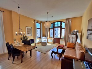 Un ampio soggiorno con tavolo da pranzo in legno, sedie nere, divano marrone e tavolino da caffè rosso. Presente una grande finestra con tende, un lampadario e vista sugli edifici esterni.