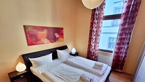 Ein ordentlich bezogenes Doppelbett mit weißen Leinen und Handtüchern, ein Kopfteil, Nachttischlampen, ein großes abstraktes Gemälde und ein Fenster mit Vorhängen.