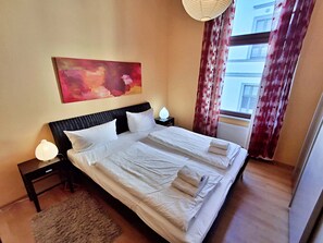 A neatly made double bed with white linens and towels, a headboard, bedside lamps, a large abstract painting, and a window with curtains.