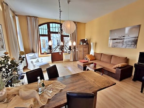 A living room with a sofa, dining table, chairs, a chandelier, and a large window.