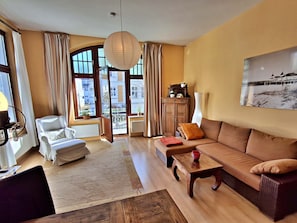 A living room with a sofa, armchair, and a table. There is a large window with curtains, a radiator, and a hanging light fixture.