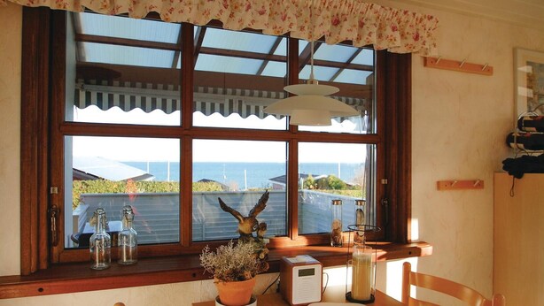 Ein Zimmer mit großem Fenster und Blick auf die Küste, einem Holztisch mit weißem Teller, einer Topfpflanze und einem Radio.