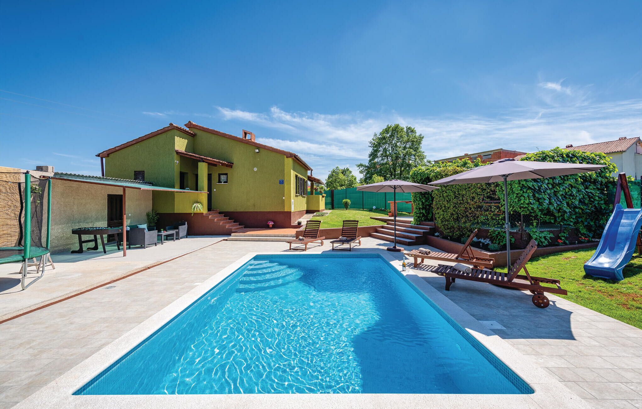 Ein Swimmingpool mit Liegestühlen und einer Rutsche, umgeben von einem Haus und Grünflächen.