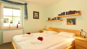 Una cama bien hecha con sábanas blancas y flores rojas, una lámpara de noche y una estantería con libros y un cuadro en la pared.