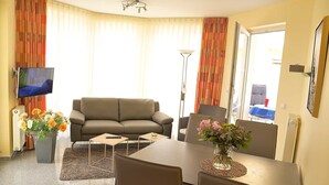 Una sala de estar con sofá, televisión, mesa de comedor con sillas y un florero con flores.