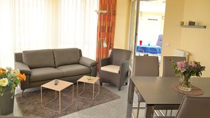 Una sala de estar con sofá, sillones, una mesa de centro y una mesa de comedor con sillas. Hay un florero con flores sobre la mesa de comedor.