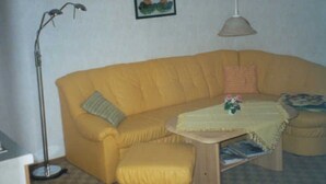 Un canapé jaune accompagné d’un pouf assorti, une petite table surmontée d’un chemin de table à motifs floraux, une lampe sur pied et un tableau encadré accroché au mur.