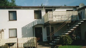 Une maison à deux étages avec un porche à l’avant, un petit jardin et une clôture en bois.