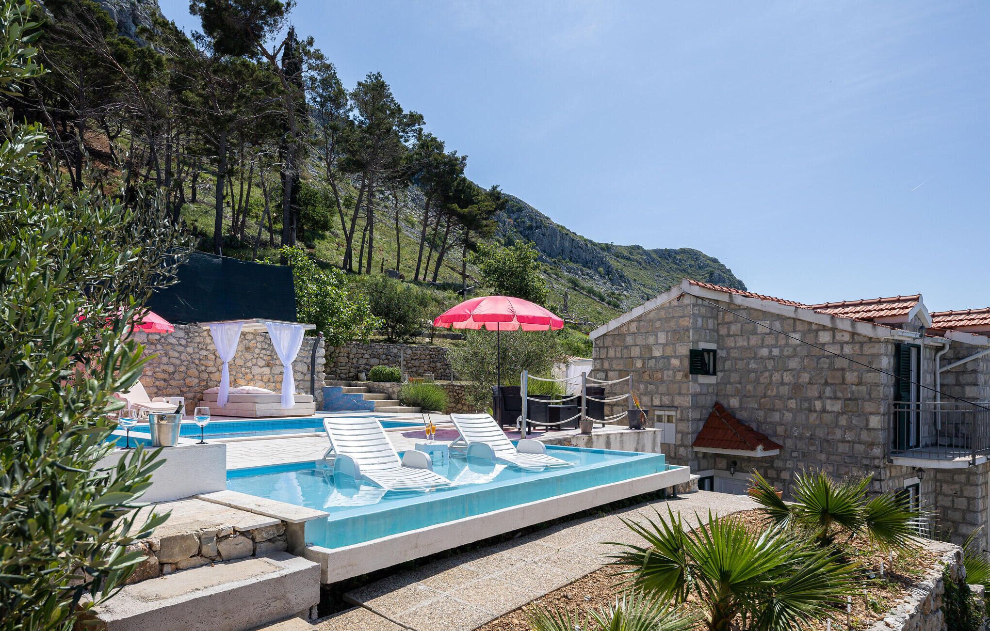 Ein Poolbereich mit Liegestühlen, ein Steinhaus und eine gebirgige Kulisse.