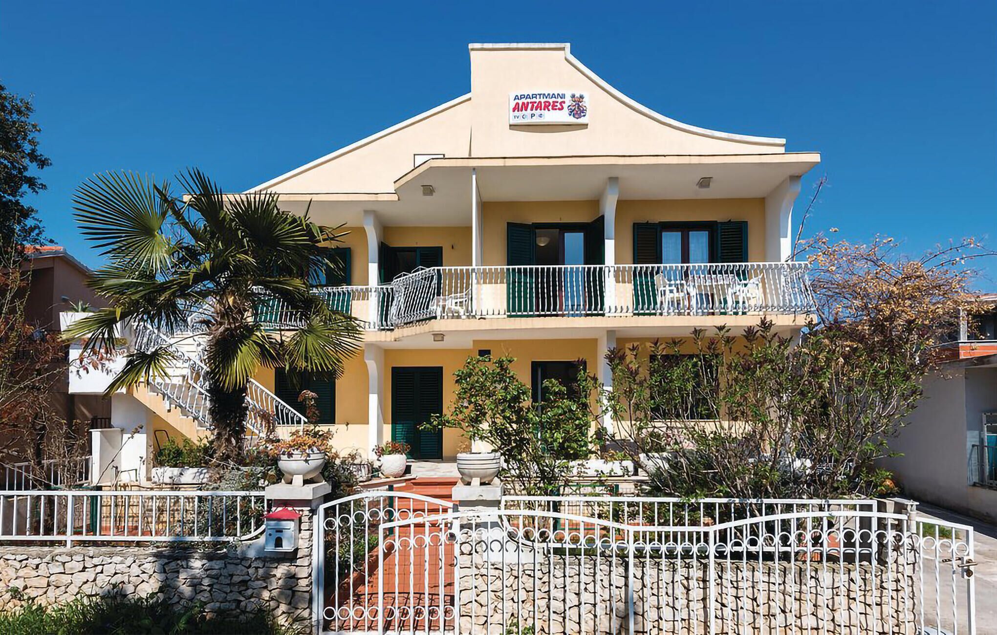 A two-story building with a sign reading "APARTMANI ANTARES." It features balconies with white railings and a stone wall in front.