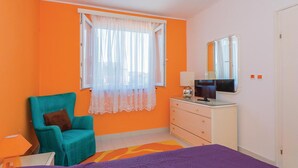Een slaapkamer met een oranje wand, een blauwgroene fauteuil, een bed met een paars deken, een dressoir met een spiegel en een raam met dunne gordijnen.