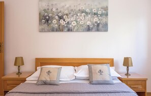 Ein ordentlich bezogenes Bett mit einer grauen Bettdecke, zwei Kissen mit aufgestickten Motiven und einem Blumenbild an der Wand.