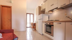 Una cocina compacta con estufa, horno y refrigerador. Hay un comedor con sillas y una mesa. Un sofá rojo se observa en primer plano.