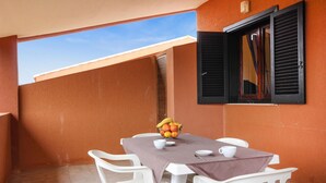 Un pequeño espacio de comedor al aire libre con una mesa dispuesta para dos personas, sillas de plástico blancas, un cuenco de fruta y una ventana con persianas negras.