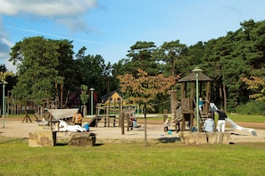 A playground with various play structures, including a slide and climbing areas, surrounded by trees and open space.