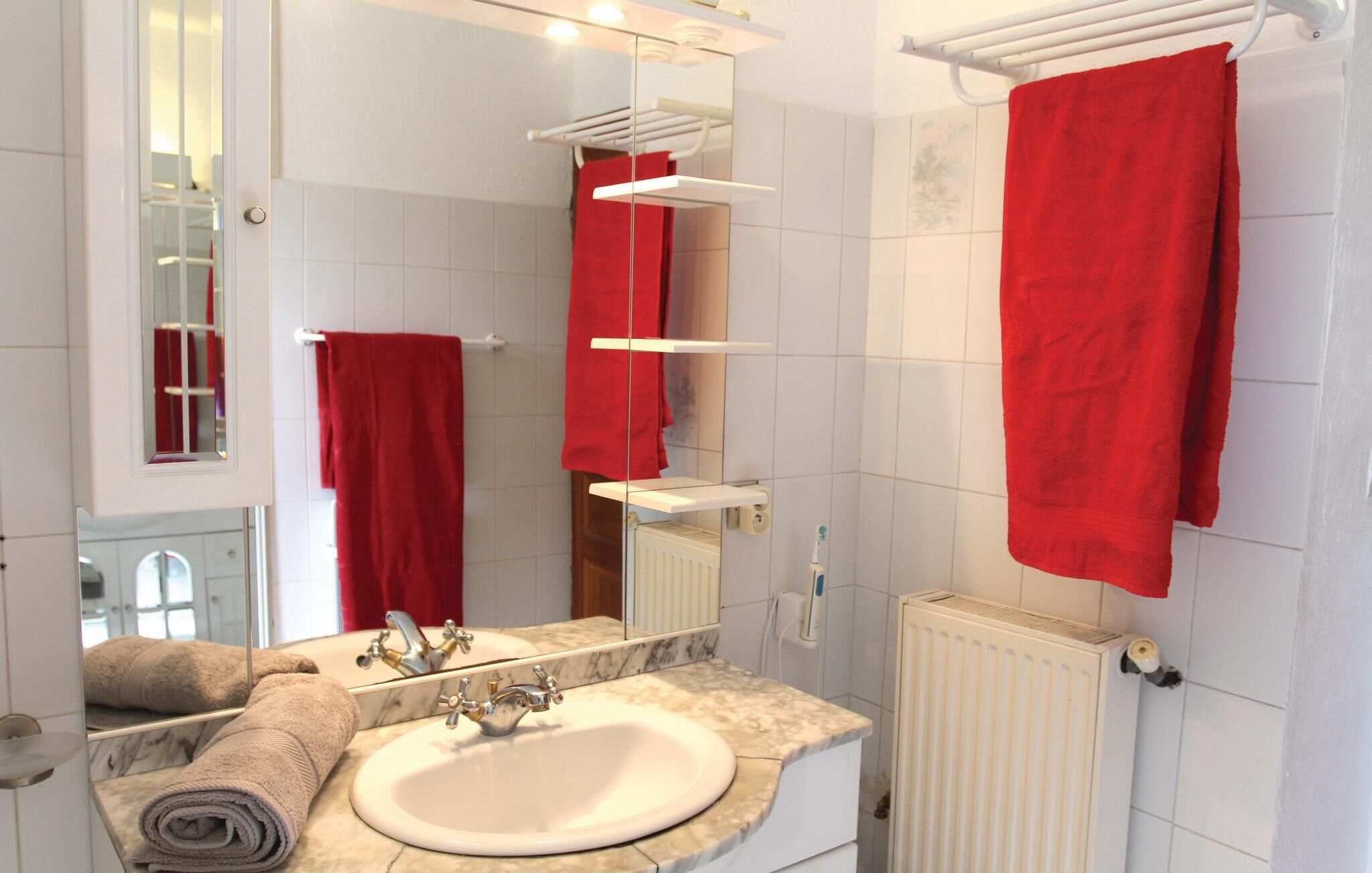 Ein Badezimmer mit einem weißen Waschbecken, einem Spiegel, einem Handtuchhalter mit roten Handtüchern und einer weißen Heizung.