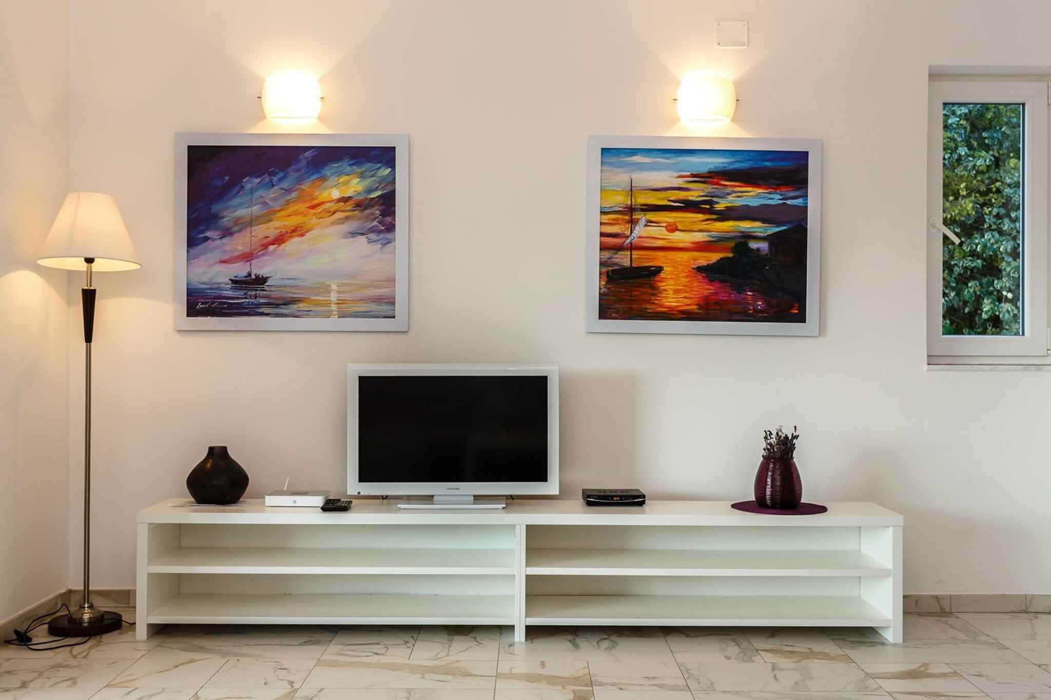 Una sala de estar moderna con un televisor de pantalla plana sobre un mueble blanco, dos cuadros en la pared que representan puestas de sol, y una lámpara de pie.