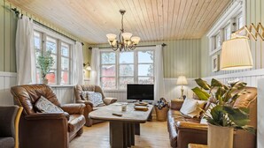 A cozy living room with leather sofas, a coffee table, and a TV. There are large windows with curtains, a chandelier, and potted plants. - Cozy home in Ebeltoft with kitchen (Ebeltoft)