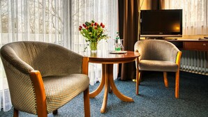 A hotel room with two armchairs, a round table with a vase of flowers, a television, and a lamp.
