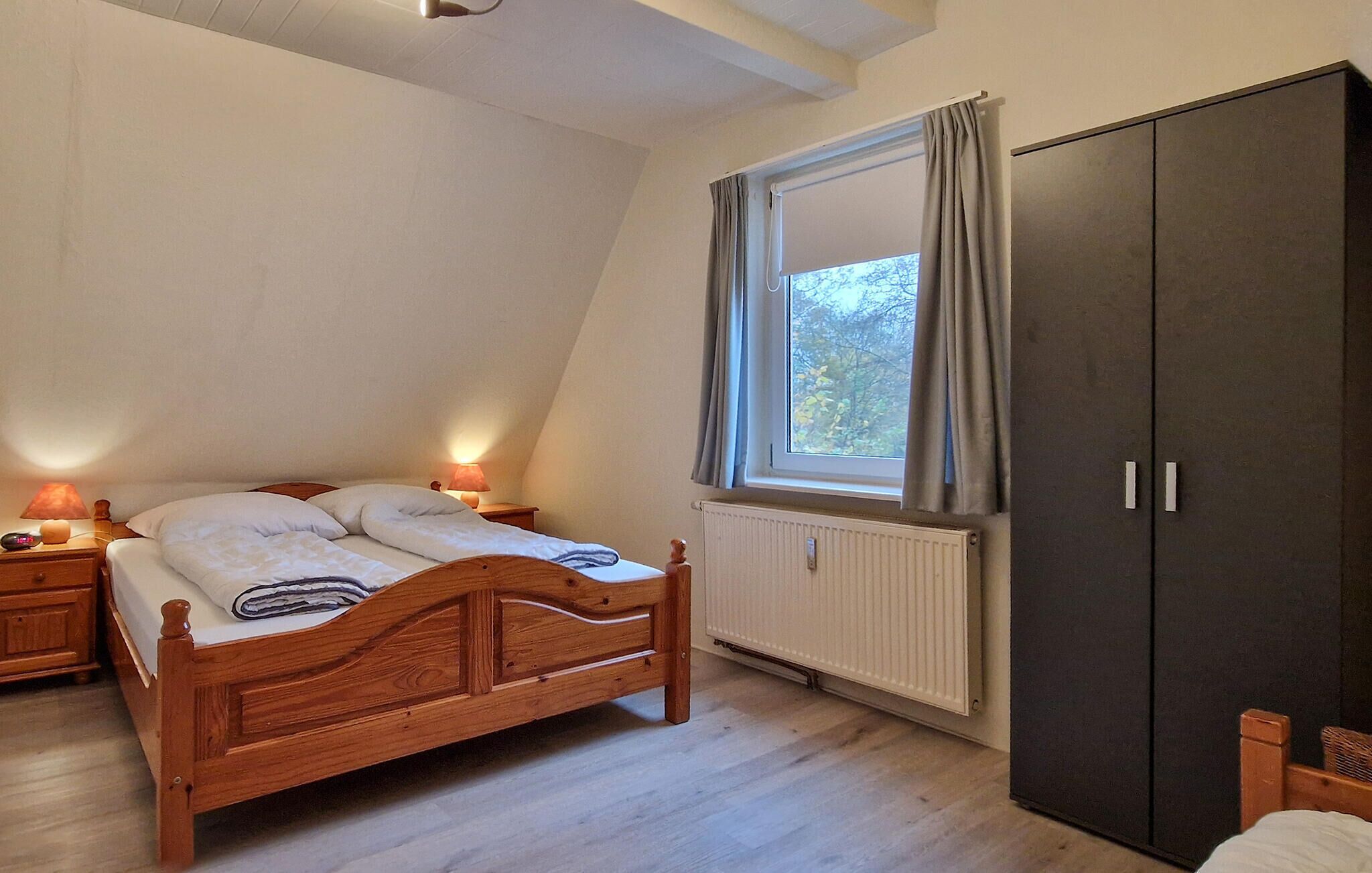 Un dormitorio con una cama de madera, una mesita de noche con lámpara, una ventana con cortinas y un armario.
