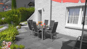 An outdoor patio with a table and chairs, a red umbrella, and a white building with a window.