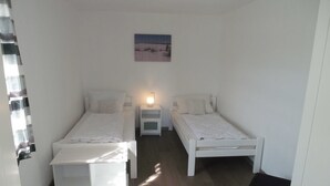 A bedroom with two single beds, a nightstand with a lamp, a small painting on the wall, and a door with a curtain. - 3-space; VO 1 - Gästehaus Vogelsang (Sierksdorf)