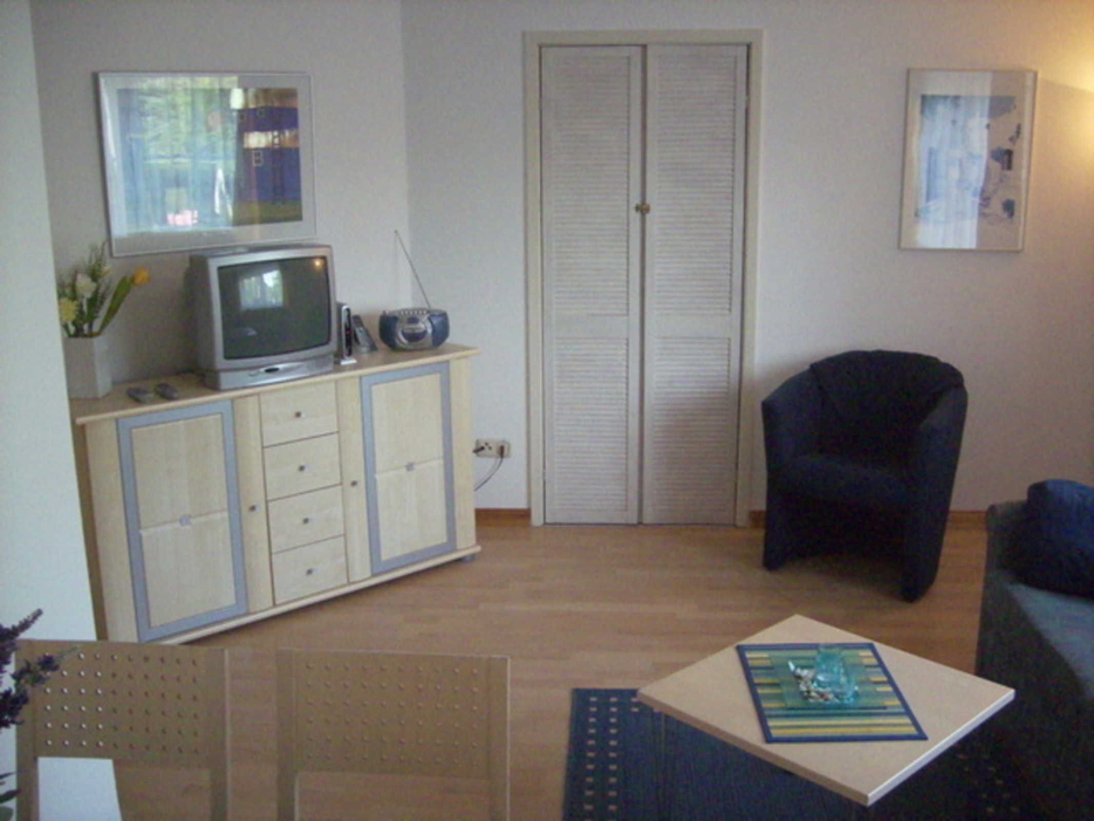 A room with a television on a cabinet, a chair, a small table with a book, and a door.