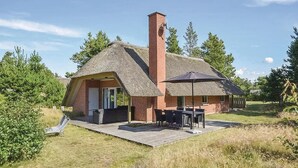 Una cabaña con techo de paja y chimenea de ladrillo, un patio con mesa y sillas, y una terraza de madera.