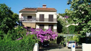 A two-story residential building with a balcony, surrounded by greenery and purple flowers. - Holiday apartment with terrace (Mali Lošinj)