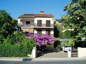 Un edificio residenziale a due piani con un balcone, circondato da vegetazione e fiori viola.