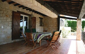 Un patio con paredes de piedra, suelo de baldosas, sillas de madera, una mesa con mantel a rayas y un farol colgante.