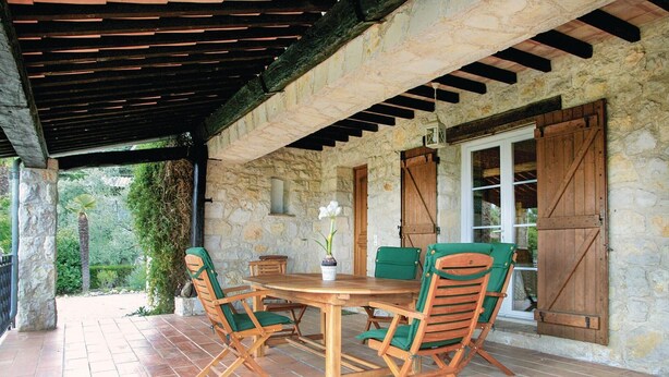 Een overdekt terras met een stenen muur, een houten tafel en groene fauteuils met kussens.