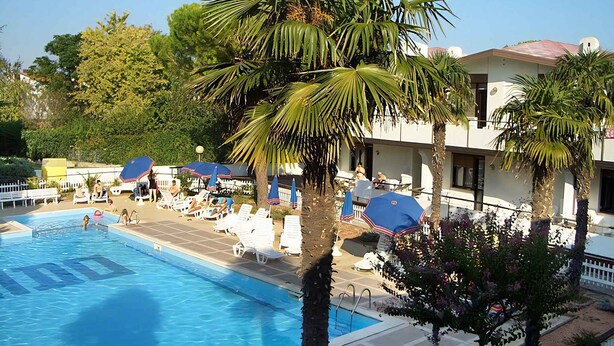 A swimming pool with lounge chairs and umbrellas, surrounded by palm trees and a building with a tiled roof.