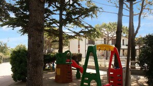 A playground with a slide and climbing structures, surrounded by trees and a building in the background.