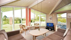 A spacious living area with a sofa set, a dining table, and chairs. A television is mounted on the wall, and there are large windows providing a view of the outdoors. - Nice home in Væggerløse with sauna (Væggerløse)