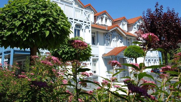 Un edificio grande y blanco con techo de tejas rojas, rodeado de abundante vegetación y plantas en flor.