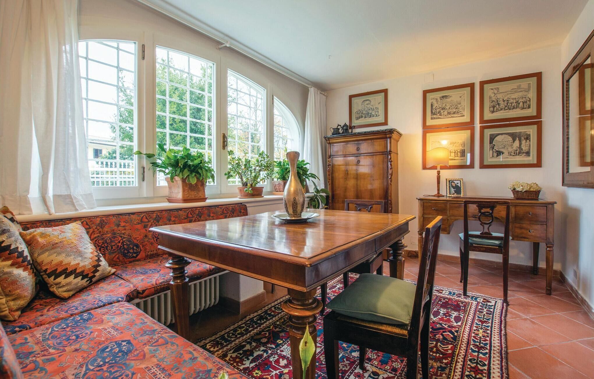 Ein gemütliches Zimmer mit einem Holzesstisch, einer Bank und einem Sofa. Große Fenster mit Vorhängen, ein Heizkörper und eine Kommode mit gerahmten Bildern an der Wand.