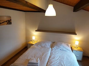 A bedroom with a bed, two bedside tables with lamps, a wooden ceiling, and a wall-mounted shelf.