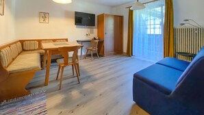 A living room with a sofa, a dining table with chairs, a TV mounted on the wall, and a window with curtains. - Ferienwohnung in Rauris (Rauris)