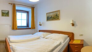 A double bed with white linens, a window with yellow curtains, a painting on the wall, and a bedside table with a lamp. - Ferienwohnung in Rauris (Rauris)