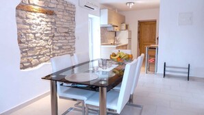 Comedor con mesa de vidrio, sillas blancas, pared de acento de piedra y cocina al fondo.