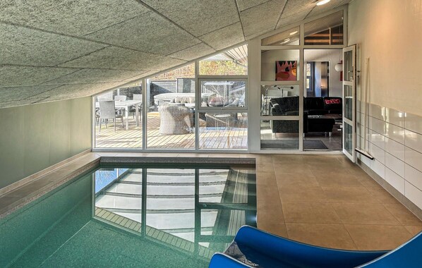 Piscina interior com paredes de vidro transparente, deck de madeira no exterior e área de estar com cadeiras azuis.