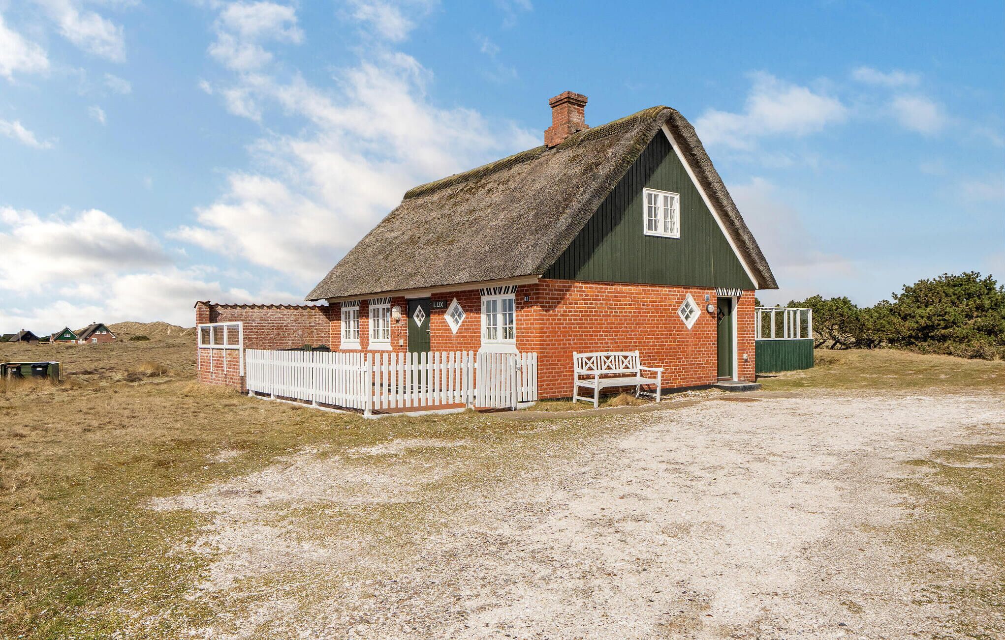 A traditional thatched-roof house with a white picket fence, a bench, and a gravel driveway.