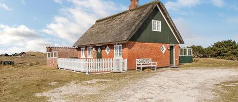 A traditional thatched-roof house with a white picket fence, a bench, and a gravel driveway.
